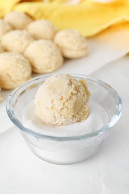 A ball of sugar cookie dough is rolled in a small bowl of sugar.