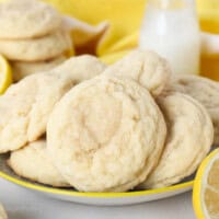 Lemon sugar cookies on a plate with a glass of milk, lemons, and more cookies in the background.