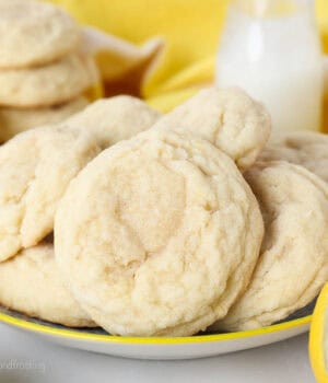 Lemon sugar cookies on a plate with a glass of milk, lemons, and more cookies in the background.