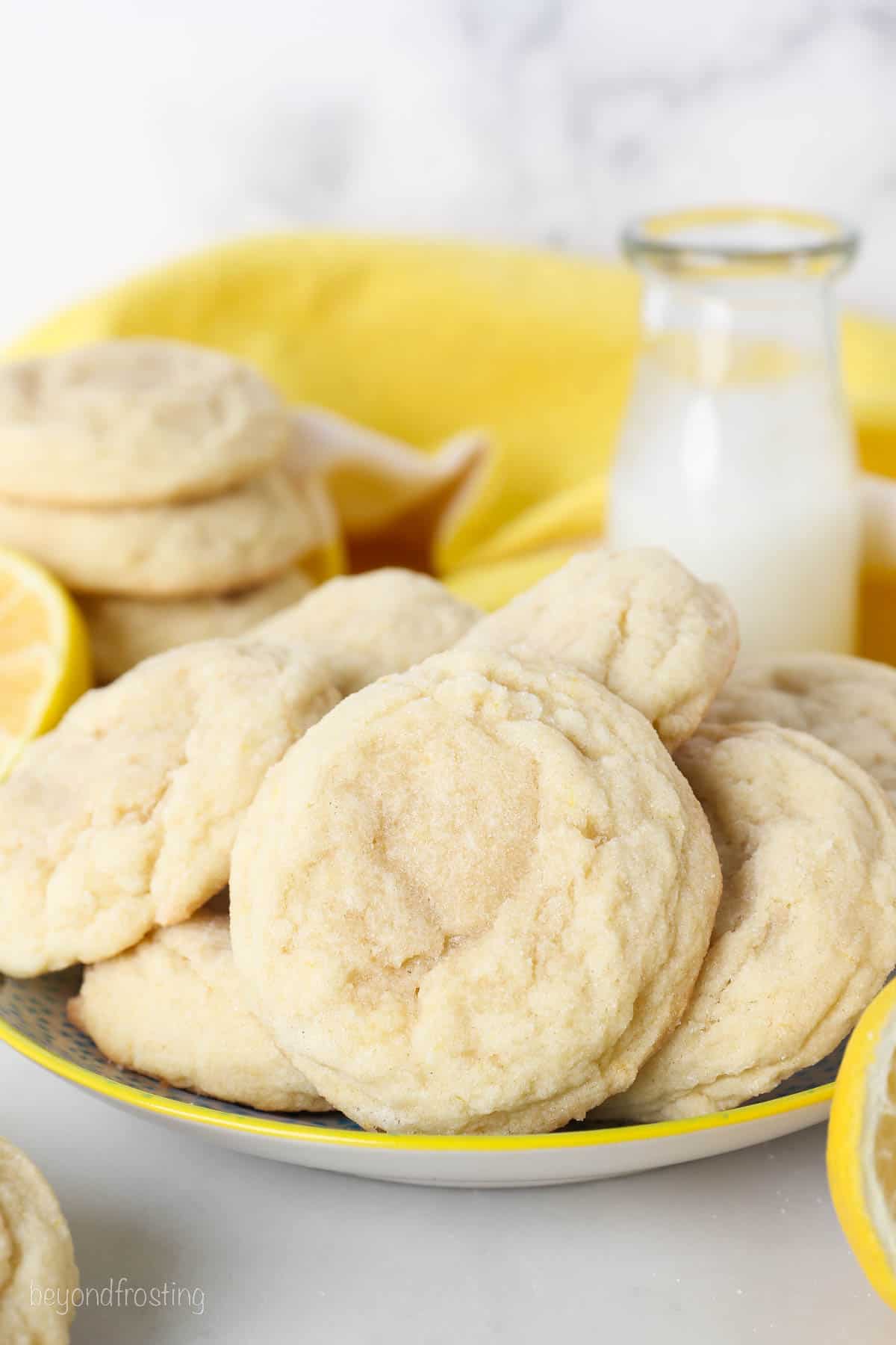 Lemon sugar cookies on a plate with a glass of milk, lemons, and more cookies in the background.