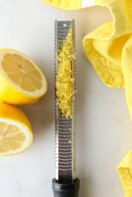 A lemon zester with zest, next to a zested lemon cut in half.