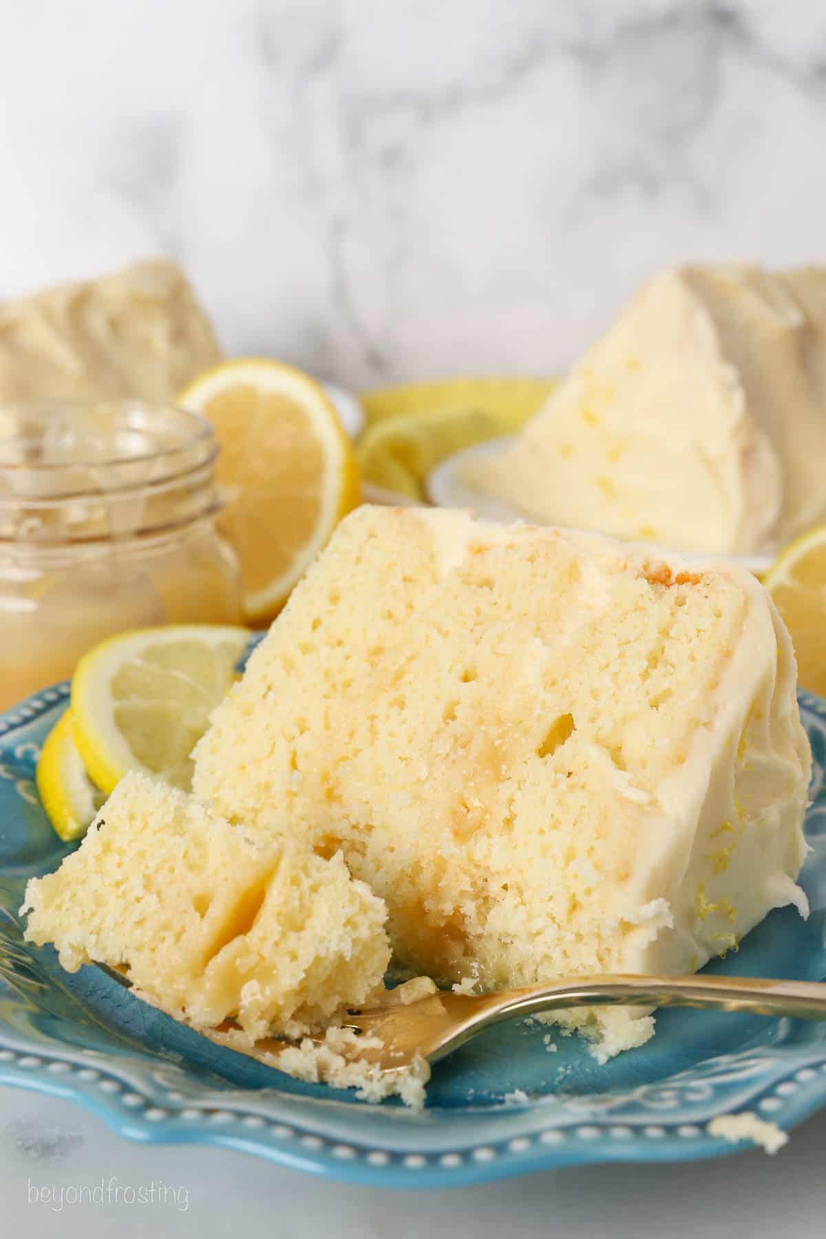 A forkful of lemon layer cake next to a slice of cake on a blue plate.