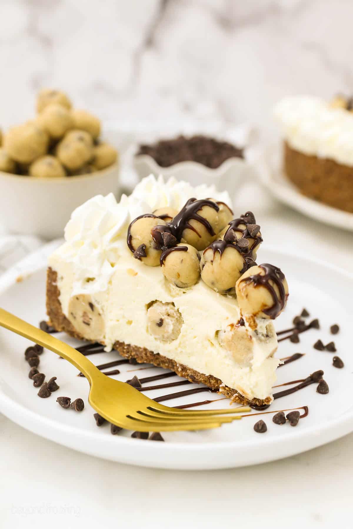 slight overhead shot of a slice of cheesecake topped with cookie dough balls on a plate next to a gold fork.