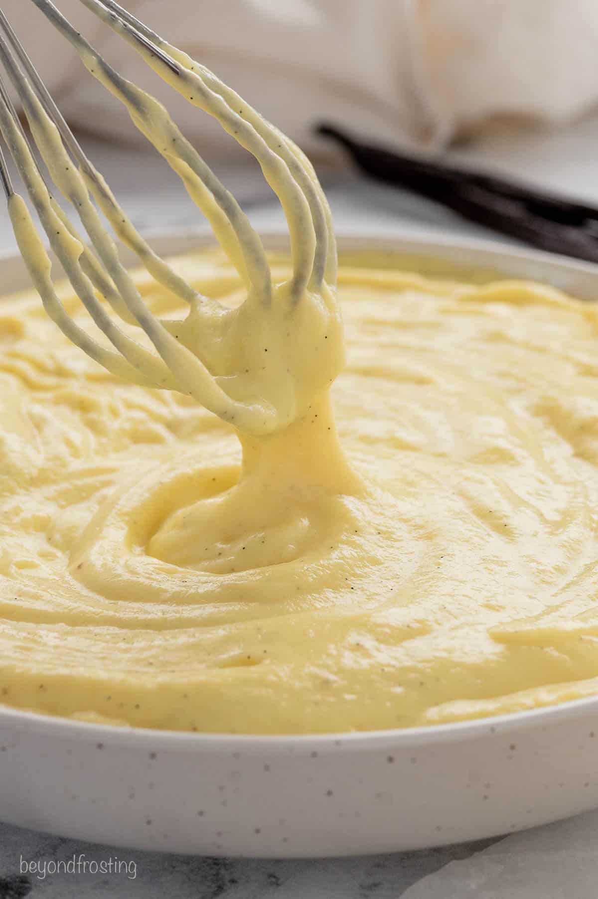 A whisk dipped into a large bowl of pastry cream.