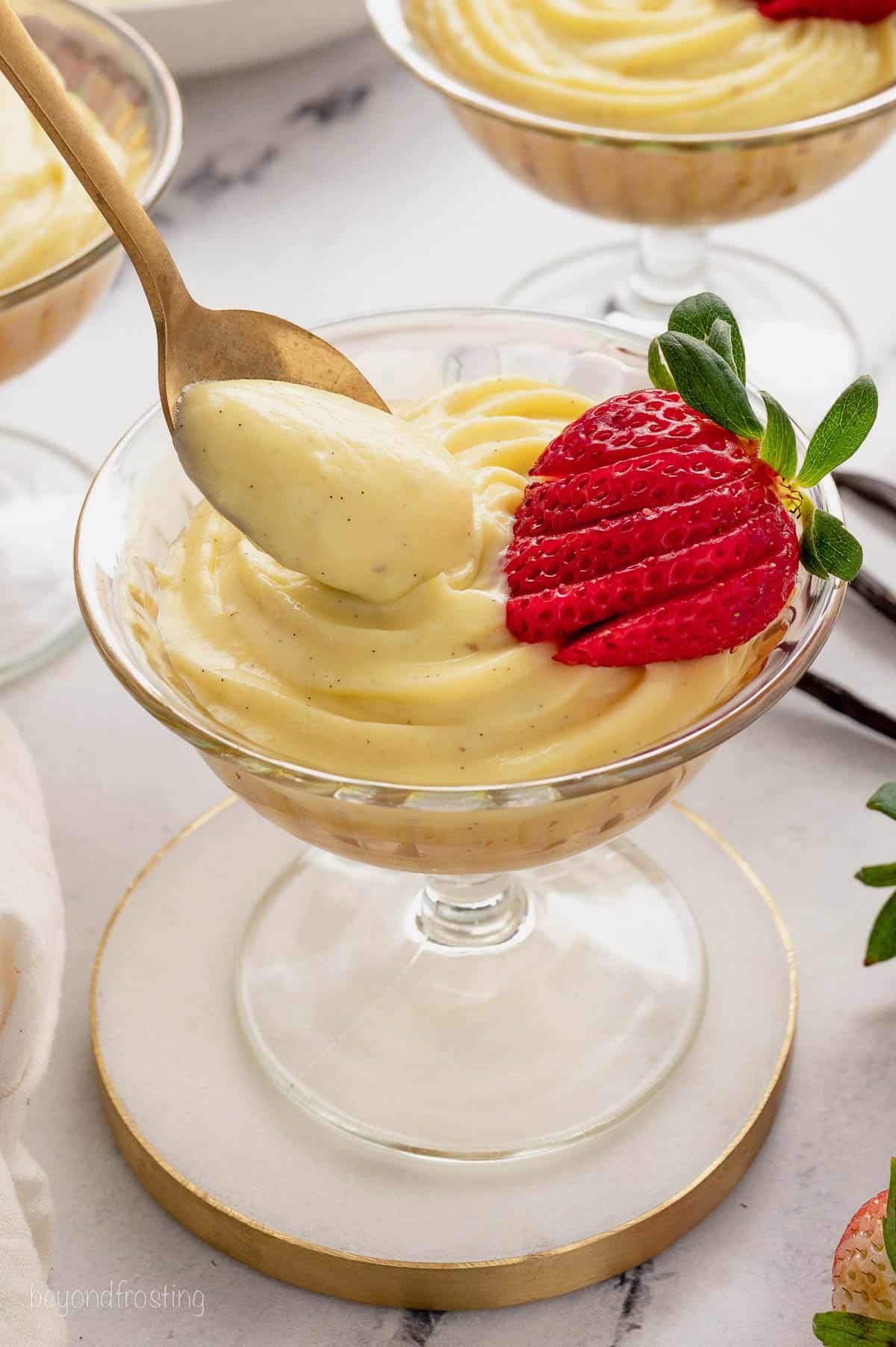 A spoon dipped into pastry cream in a coup glass, garnished with a sliced strawberry.