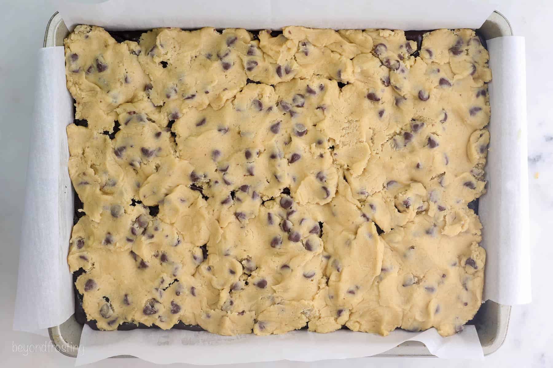Chocolate chip cookie dough layered overtop brownies and Oreos in a large baking pan.