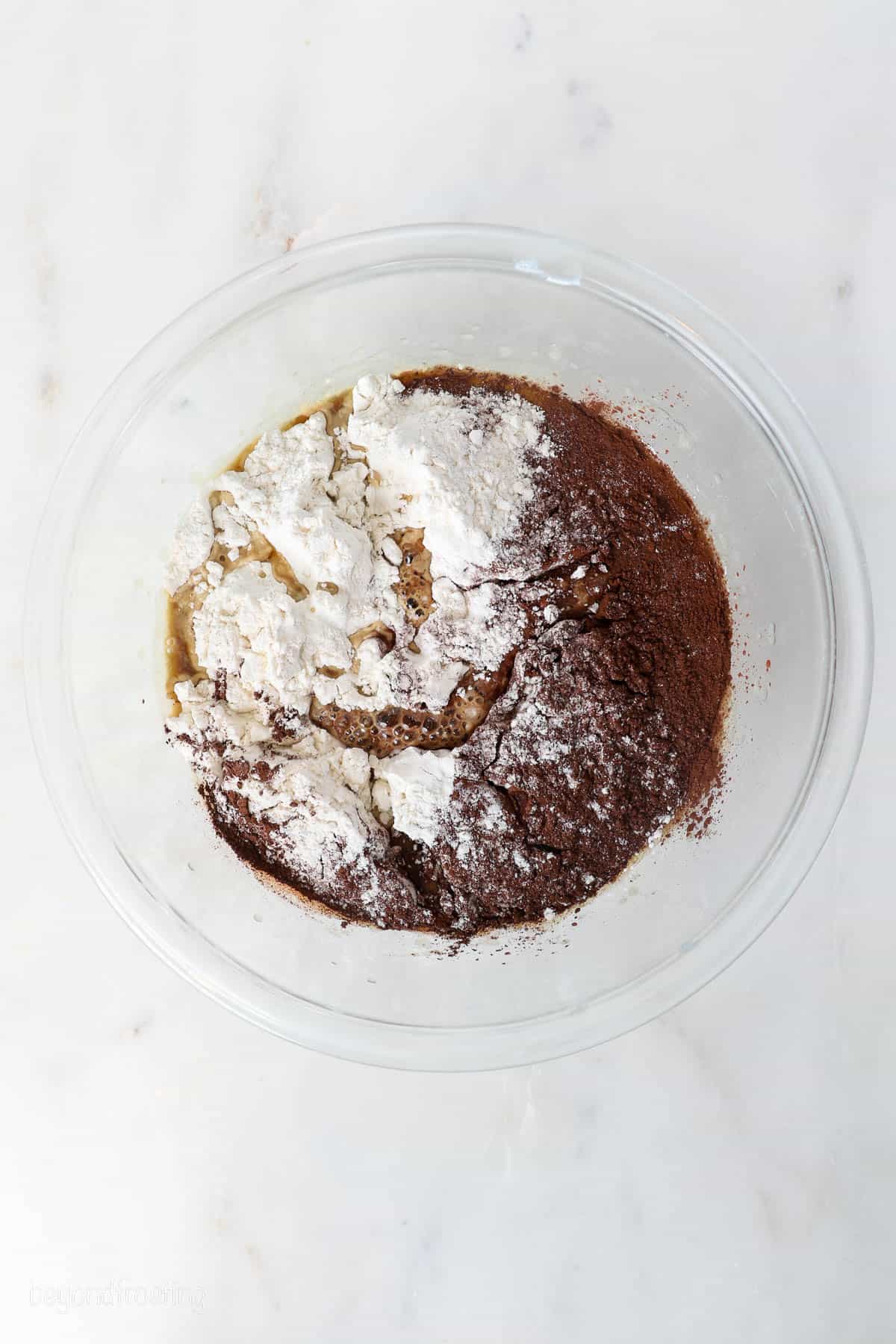 This chocolate mousse cake recipe is easy to make, with layers of moist chocolate cake, fluffy chocolate mousse, and creamy chocolate ganache. Flour and cocoa powder added to wet batter ingredients in a glass mixing bowl.