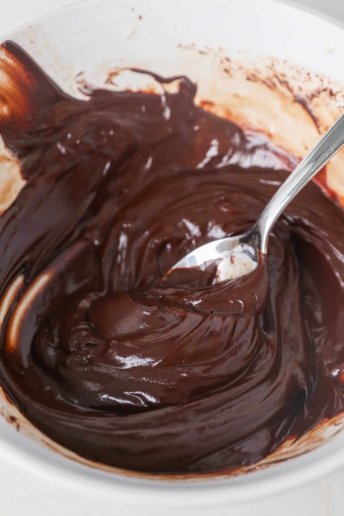 This chocolate mousse cake recipe is easy to make, with layers of moist chocolate cake, fluffy chocolate mousse, and creamy chocolate ganache. Melted chocolate ganache in a bowl with a spoon.
