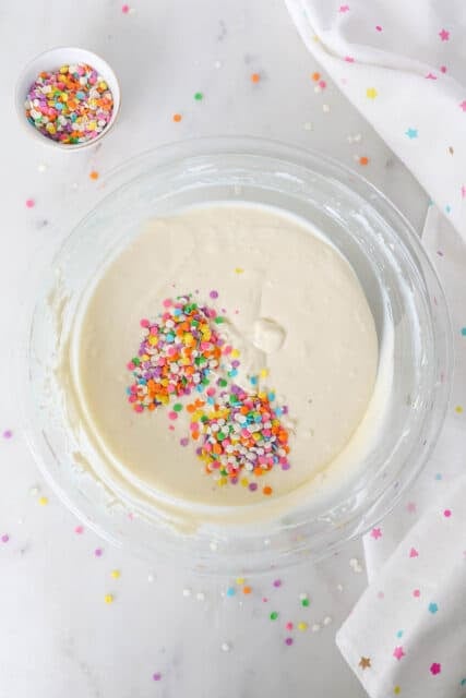 Rainbow sprinkles added to a bowl of vanilla cheesecake batter.