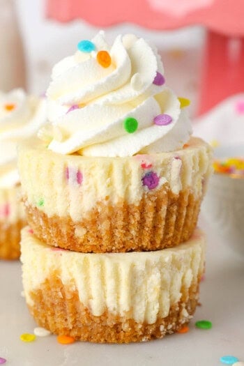 Two mini funfetti cheesecakes stacked on top of one another, with the top cheesecake decorated with a swirl of whipped cream and sprinkles.
