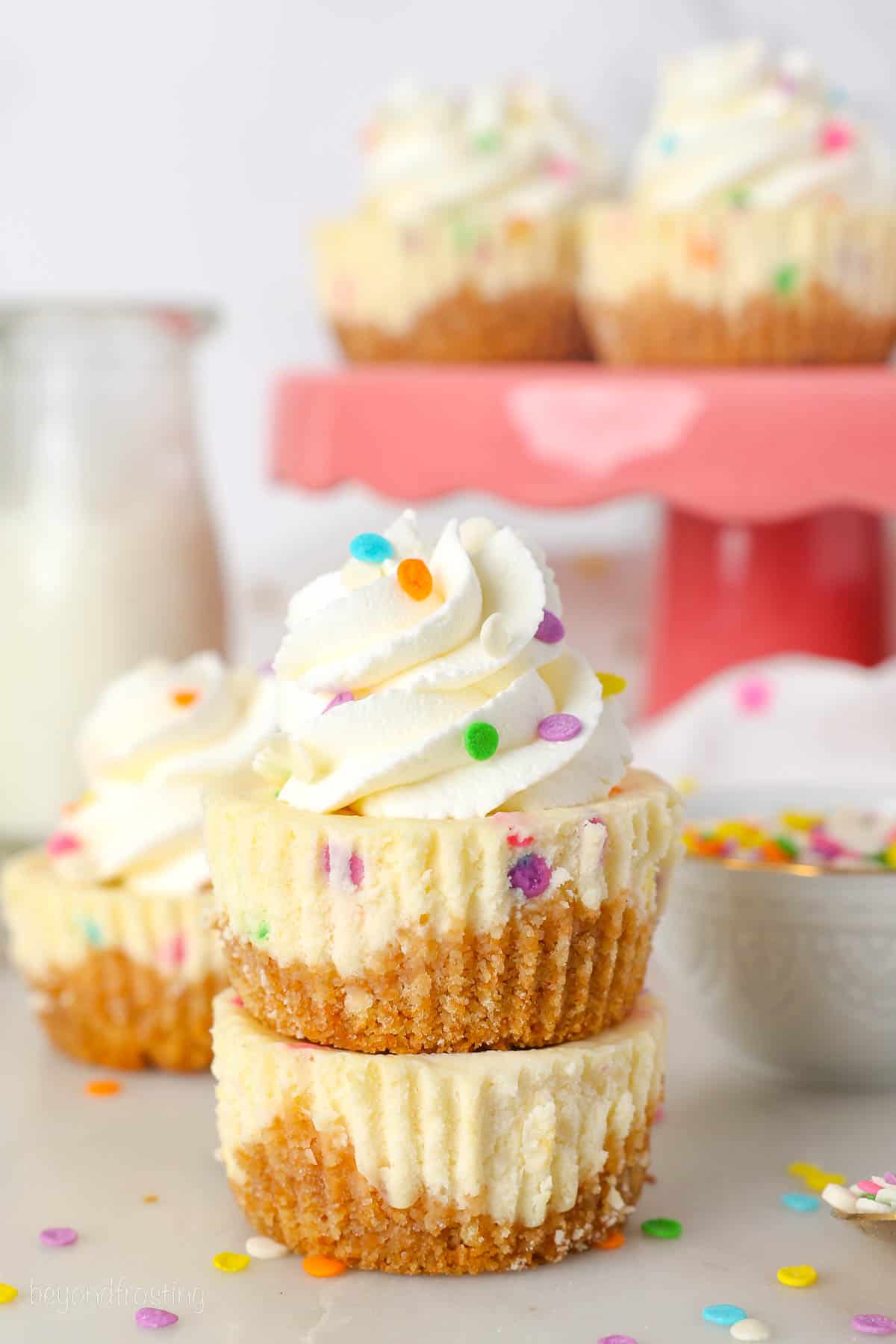 Two mini funfetti cheesecakes stacked on top of one another with a swirl of whipped cream and sprinkles, plus more cheesecakes on a cake stand in the background.