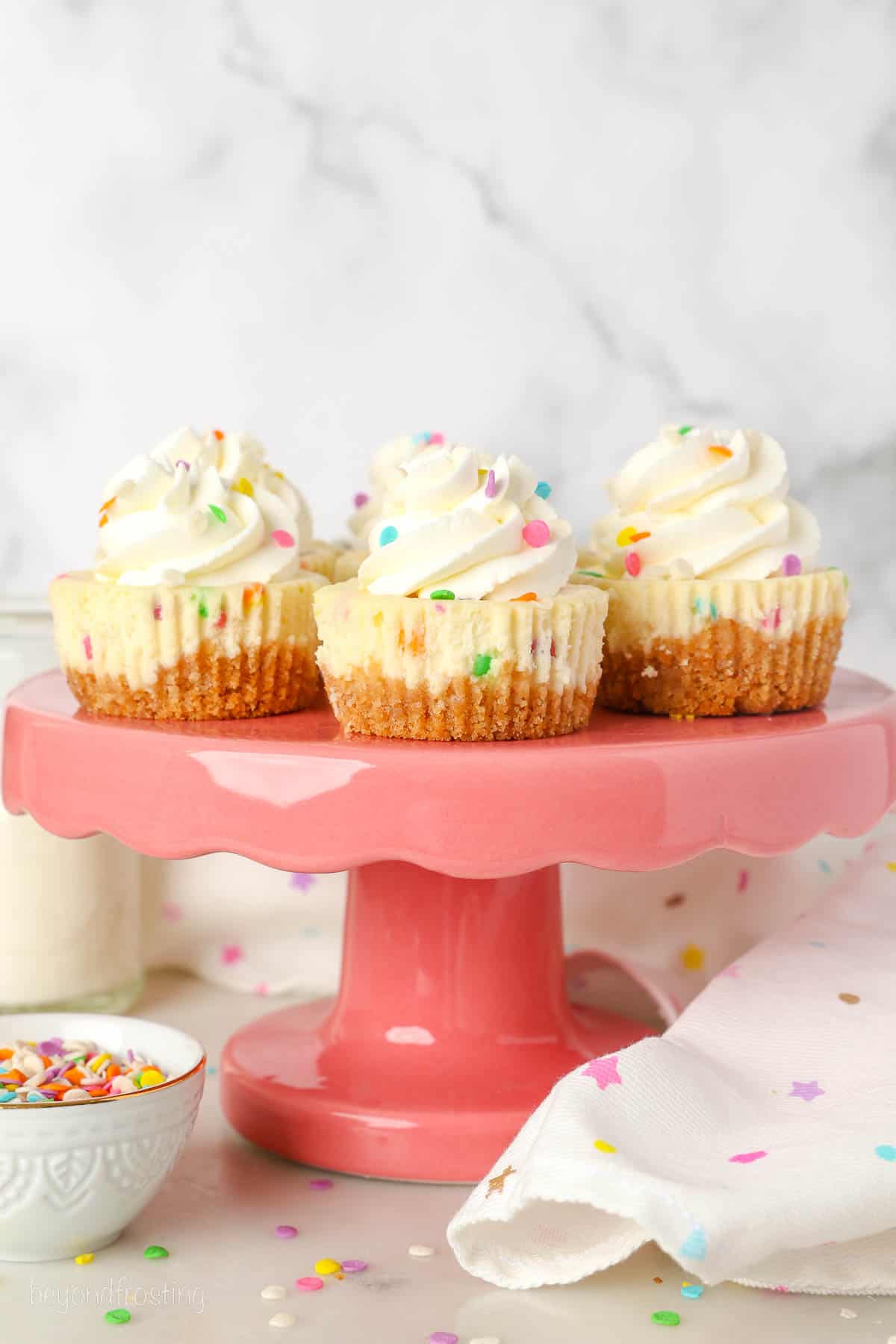 Mini funfetti cheescakes on a pink cake stand, next to a white dishcloth and a bowl of sprinkles.