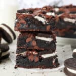 A stack of three Oreo brownies surrounded by Oreo cookies, with more brownies in the background.