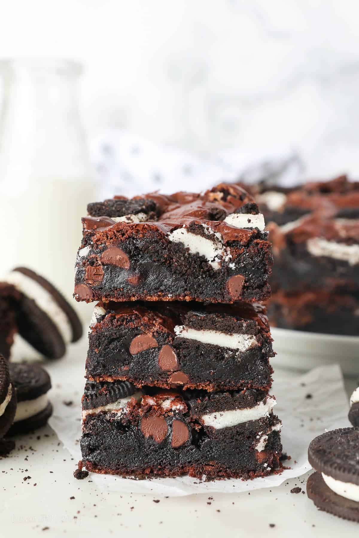 A stack of three Oreo brownies surrounded by Oreo cookies, with more brownies in the background.