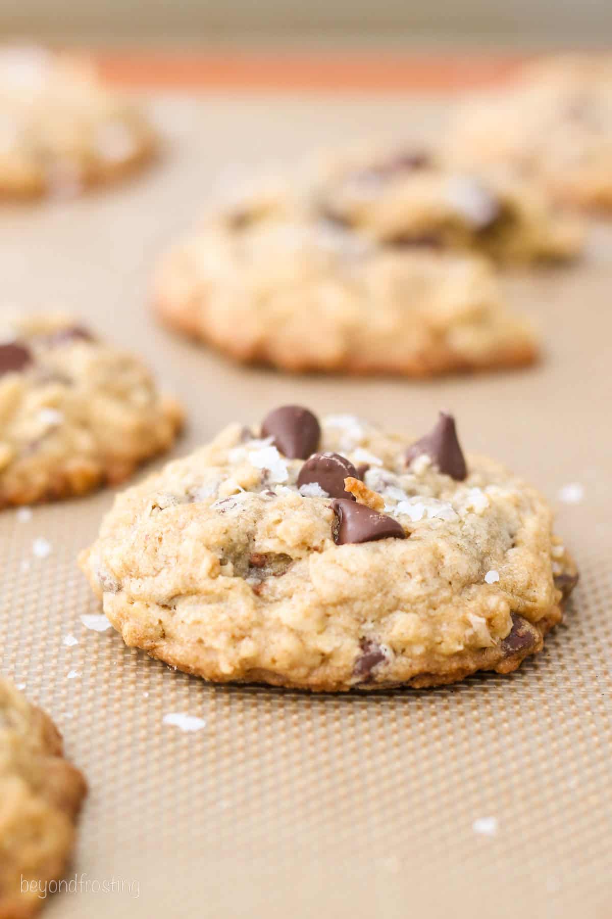 An example of a thick chocolate chip oatmeal cookie prepared with cold butter on a silicone lined baking sheet, with more cookies in the background.
