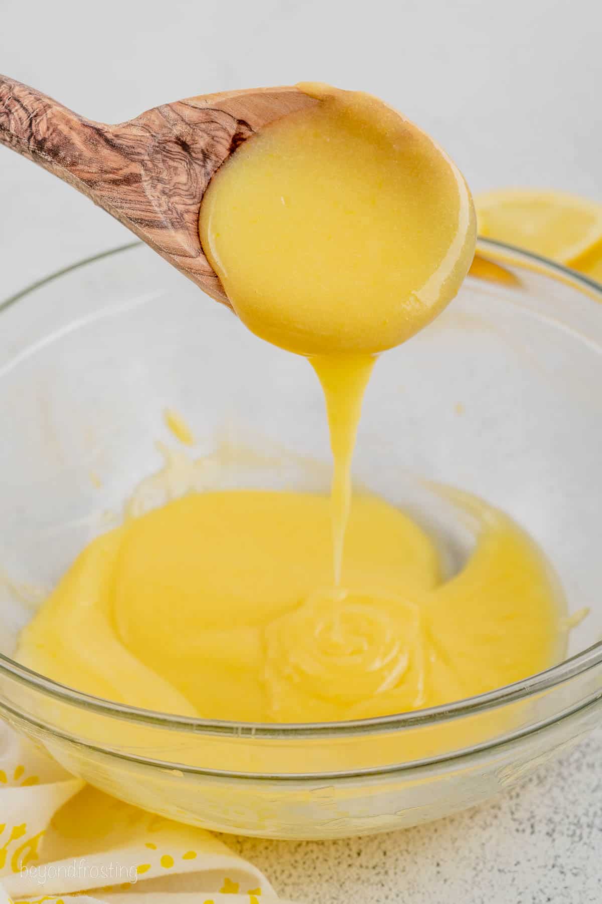 A wooden spoon dipped into lemon curd and held above a glass bowl.