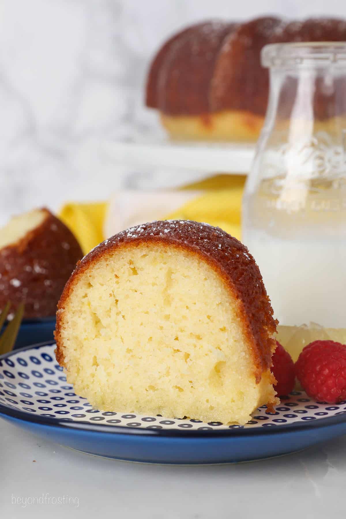 A slice of lemon drizzle cake on a plate next to fresh raspberries, with a jug of milk and a bundt cake on a cake stand in the background.