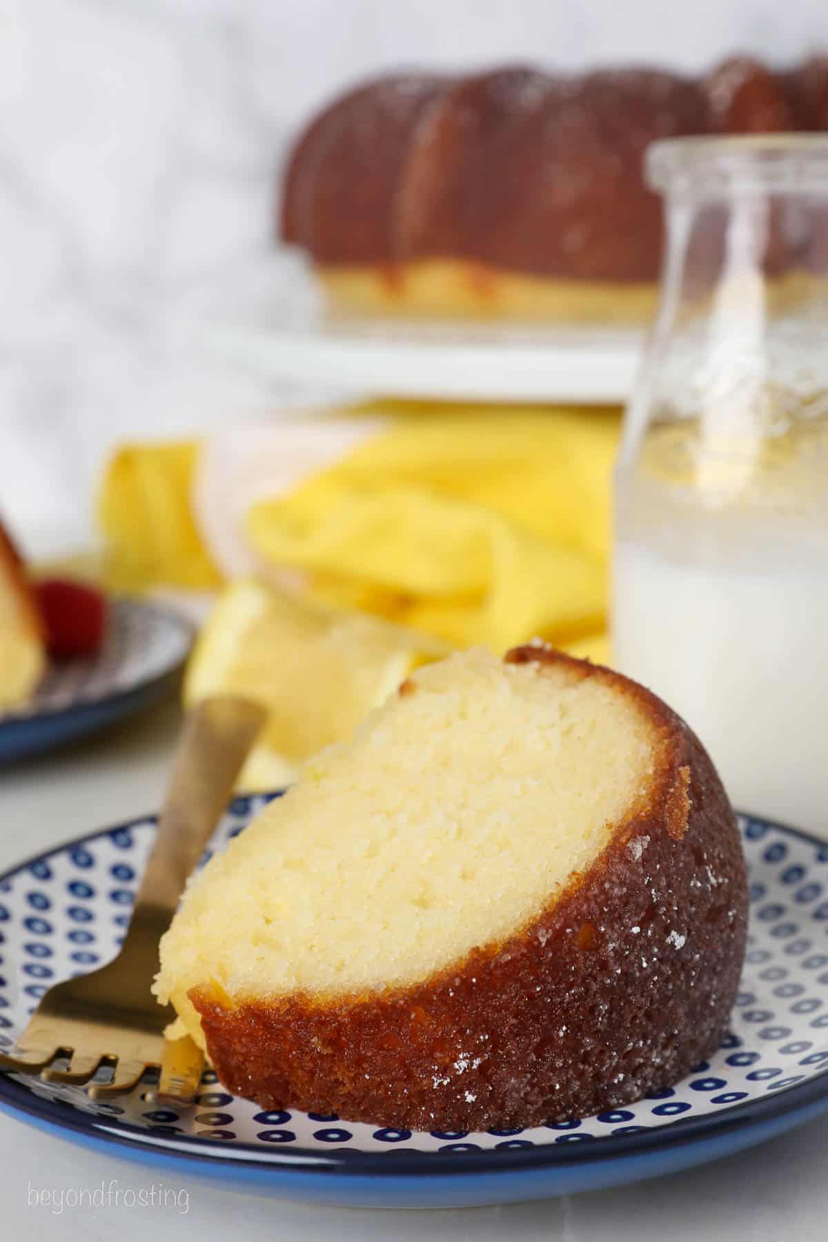 A slice of lemon drizzle cake on a plate next to a fork, with a jug of milk and a bundt cake on a cake stand in the background.