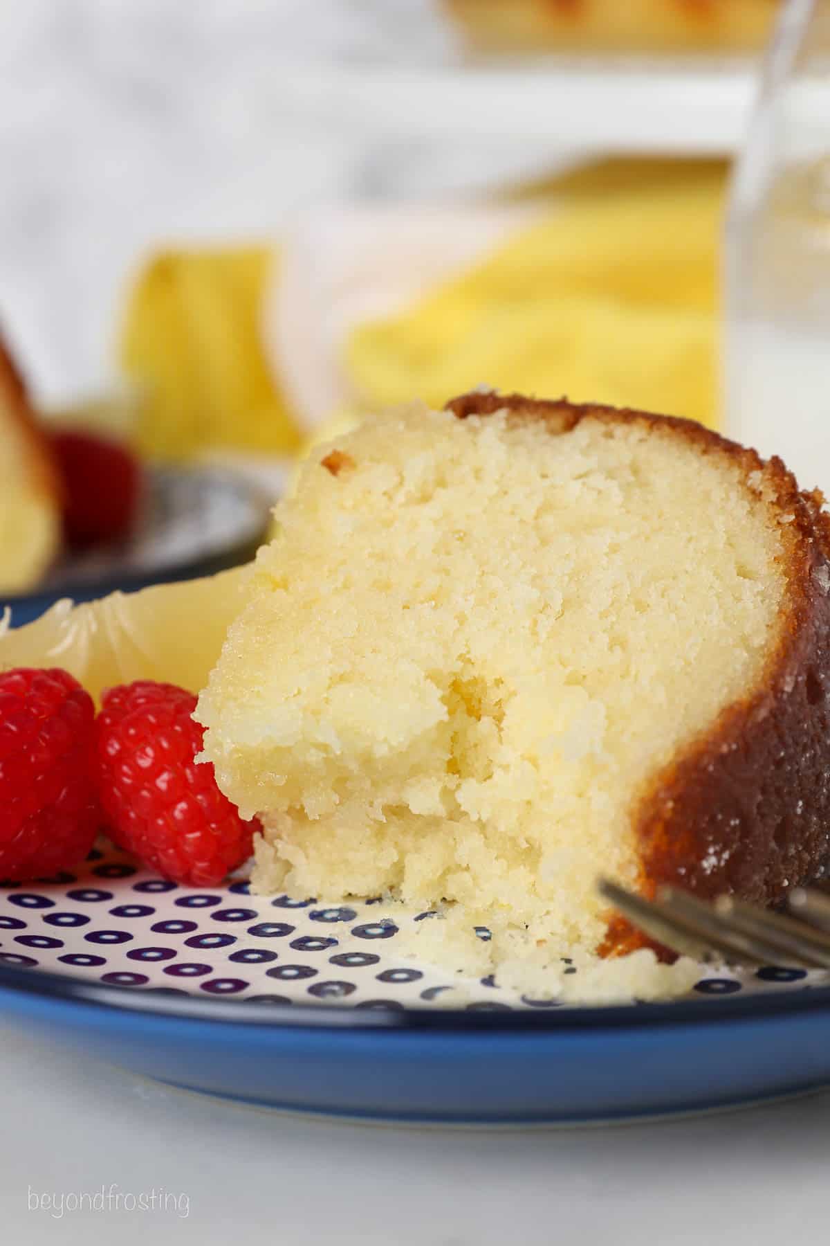 A slice of lemon drizzle cake on a plate next to fresh raspberries, with a forkful missing.