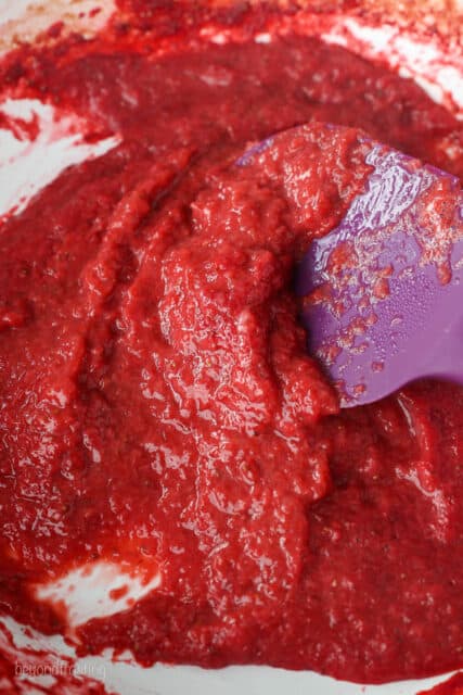 A purple spatula stirring pureed strawberries as they reduce in a skillet.