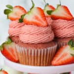 Three strawberry cupcakes with strawberry frosting on a light pink cake stand, garnished with a strawberry and surrounded by more cupcakes.