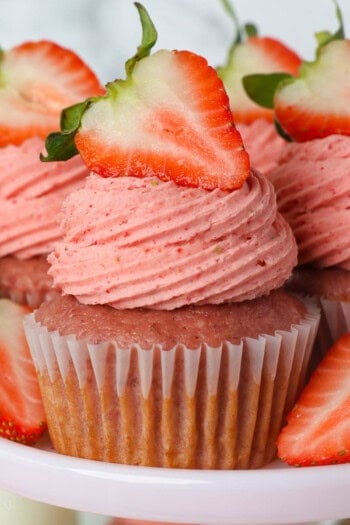 Three strawberry cupcakes with strawberry frosting on a light pink cake stand, garnished with a strawberry and surrounded by more cupcakes.