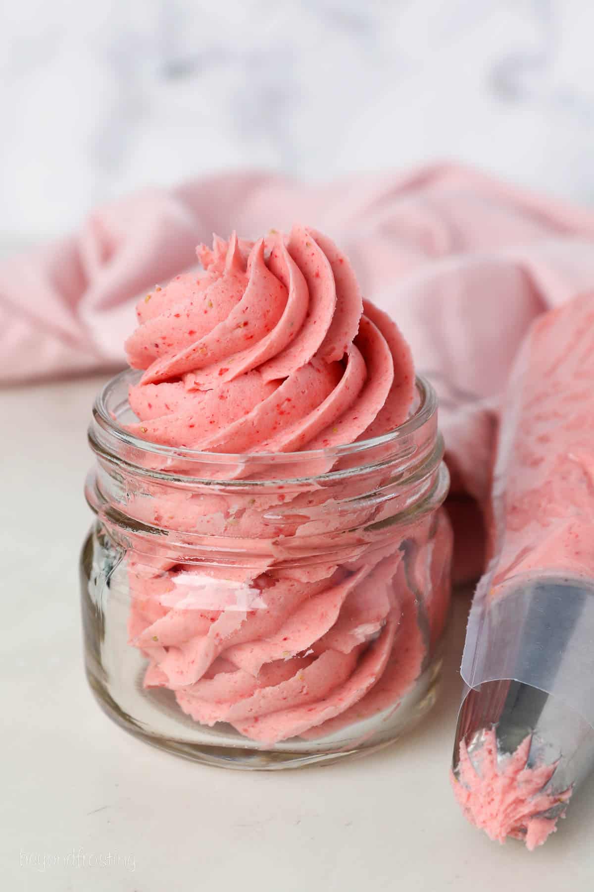 Strawberry frosting swirled inside a glass jar next to a piping bag.
