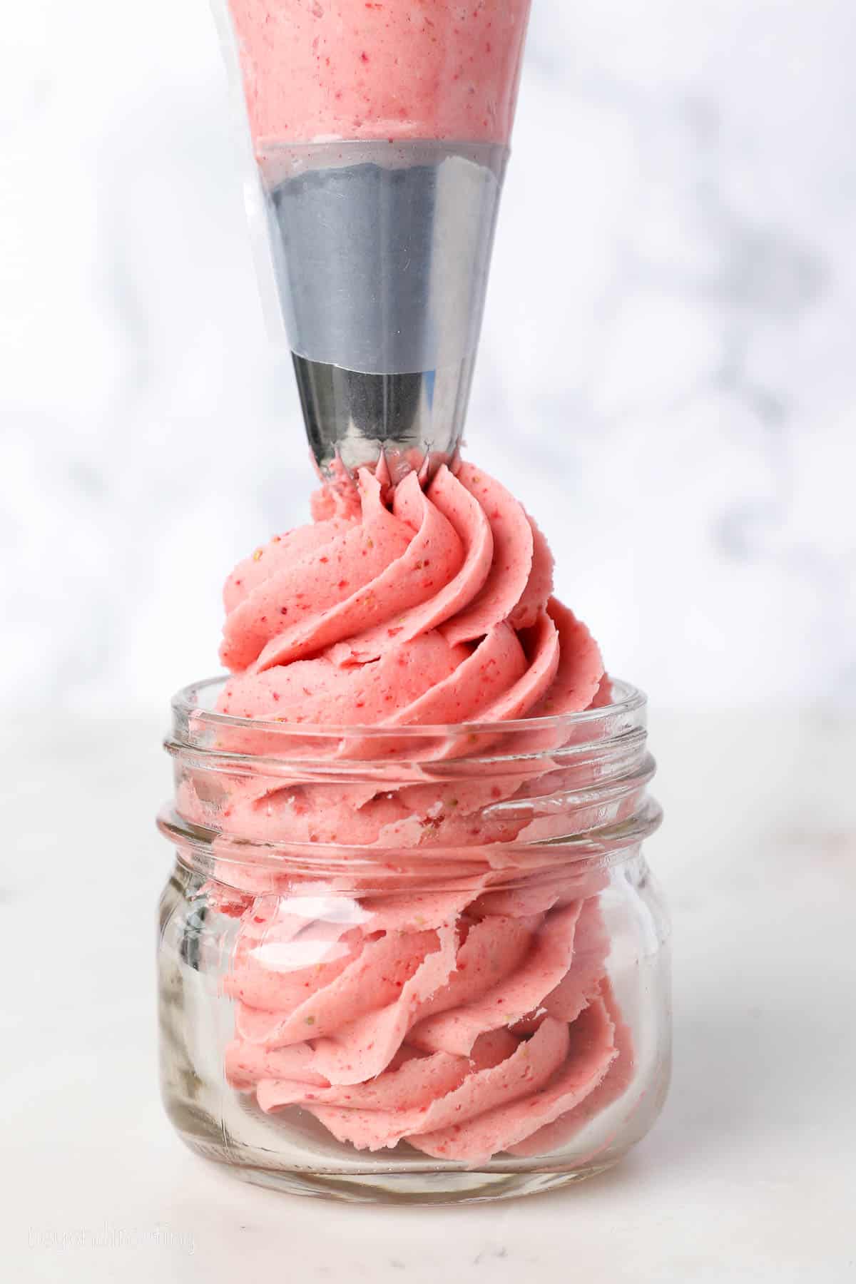 A piping tip swirls strawberry frosting into a glass jar.