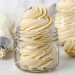 A glass jar filled with a piped swirl of brown butter frosting, with a second jar and a piping bag in the background.