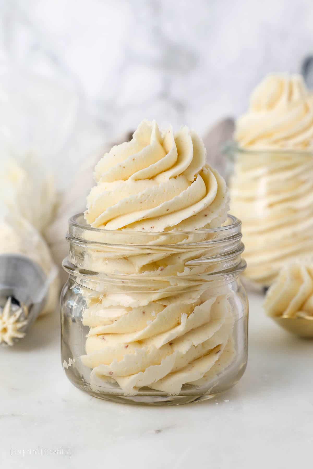 A glass jar filled with a piped swirl of brown butter frosting, with a second jar and a piping bag in the background.