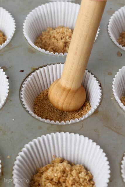 The end of a wooden spoon is used to press the graham cracker crust into the bottom of a cupcake liner in a lined cupcake pan.