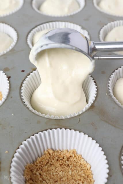 A scoop of lemon cheesecake filling is added to a cupcake liner in a lined cupcake pan.