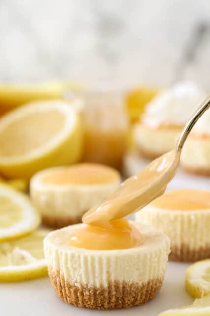 A spoon drops a dollop of lemon curd onto the top of a mini lemon cheesecake.