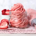 Strawberry frosting swirled inside a glass jar on top of a pink napkin with a piping bag and freeze-dried stawberries next to it