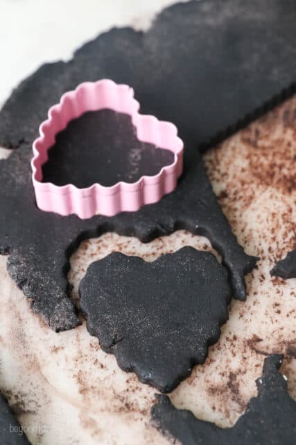 A heart-shaped cookie cutter is used to cut shapes from chocolate sugar cookie dough.