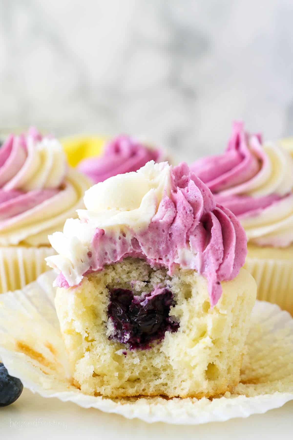 A cupcake with a bite missing to show the blueberry filling inside. It's frosted with a vanilla and blueberry swirled frosting
