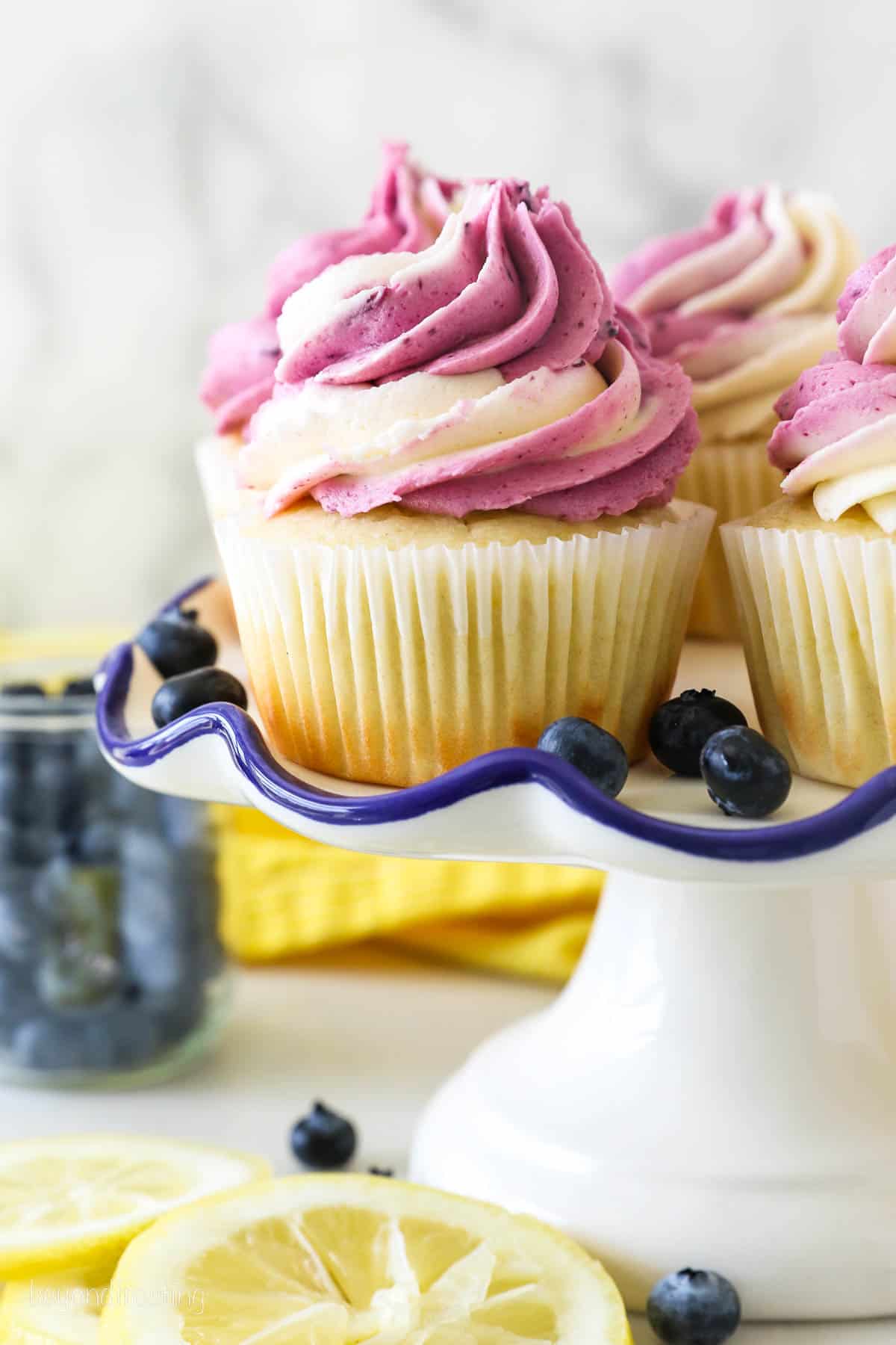 A white cake stand with lemon cupcakes frosted with a swirl of vanilla and blueberry frosting. The cake stand is surrounded by fresh cut lemons and blueberries
