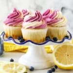 A white ruffled edge cake stand with lemon cupcakes frosted with a swirl of vanilla and blueberry frosting. The cake stand is surrounded by fresh cut lemons and blueberries