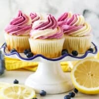 A white ruffled edge cake stand with lemon cupcakes frosted with a swirl of vanilla and blueberry frosting. The cake stand is surrounded by fresh cut lemons and blueberries
