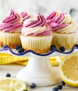 A white ruffled edge cake stand with lemon cupcakes frosted with a swirl of vanilla and blueberry frosting. The cake stand is surrounded by fresh cut lemons and blueberries