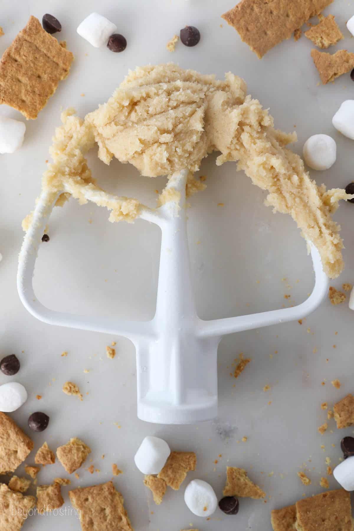 S'mores cookie dough on the end of a stand mixer attachment, resting on a countertop surrounded by scattered crushed graham crackers, marshmallows, and chocolate chips.