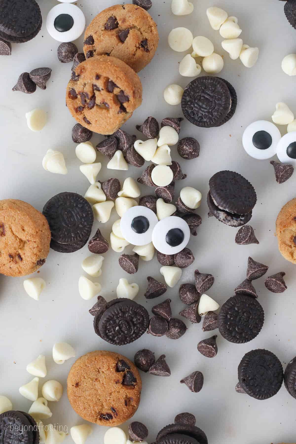 Overhead view of mini Oreos, mini Chips Ahoy cookies, white chocolate chips, chocolate chips, and candy eyes scattered on a white countertop.