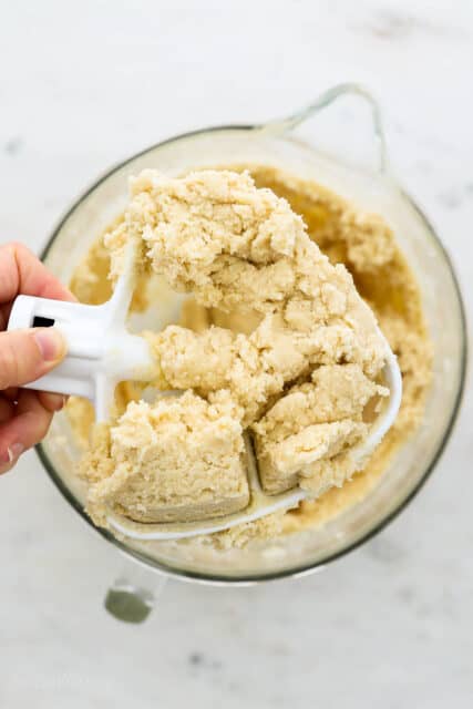 A hand holding a stand mixer attachment with snickerdoodle cookie dough clumped on the end, over a mixing bowl.