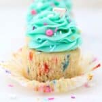 Close up of a partially unwrapped funfetti cupcake frosted with a teal buttercream swirl and sprinkles.