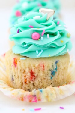 Close up of a partially unwrapped funfetti cupcake frosted with a teal buttercream swirl and sprinkles.