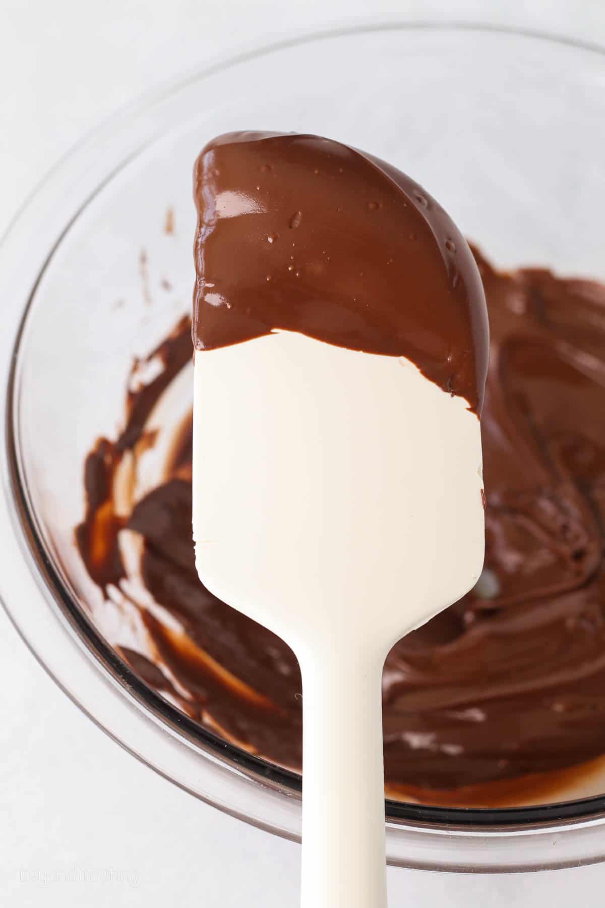 Melted chocolate on a white rubber spatula, held over a bowl of melted chocolate in the background.