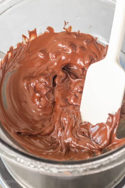 A spatula stirs partially melted chocolate chips in the glass bowl of a double boiler.