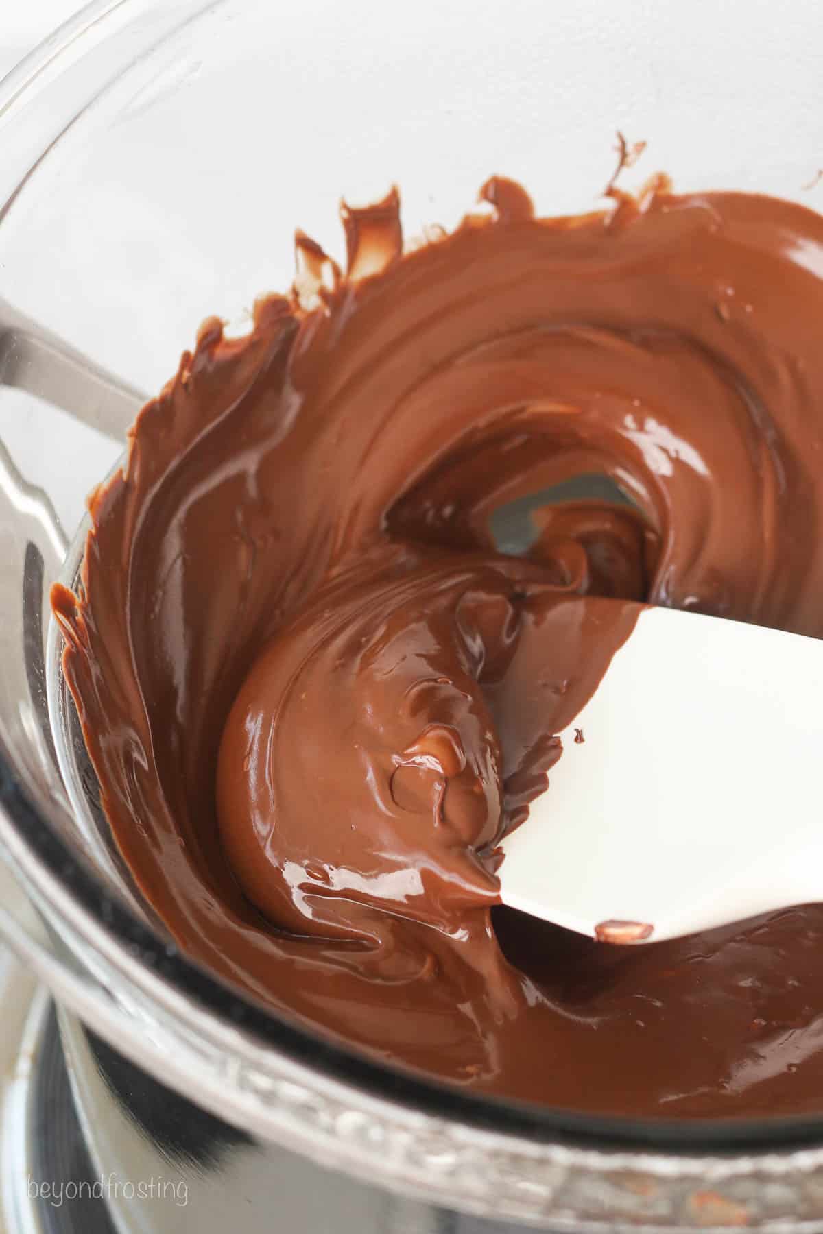 A spatula stirs partially melted chocolate chips in the glass bowl of a double boiler.