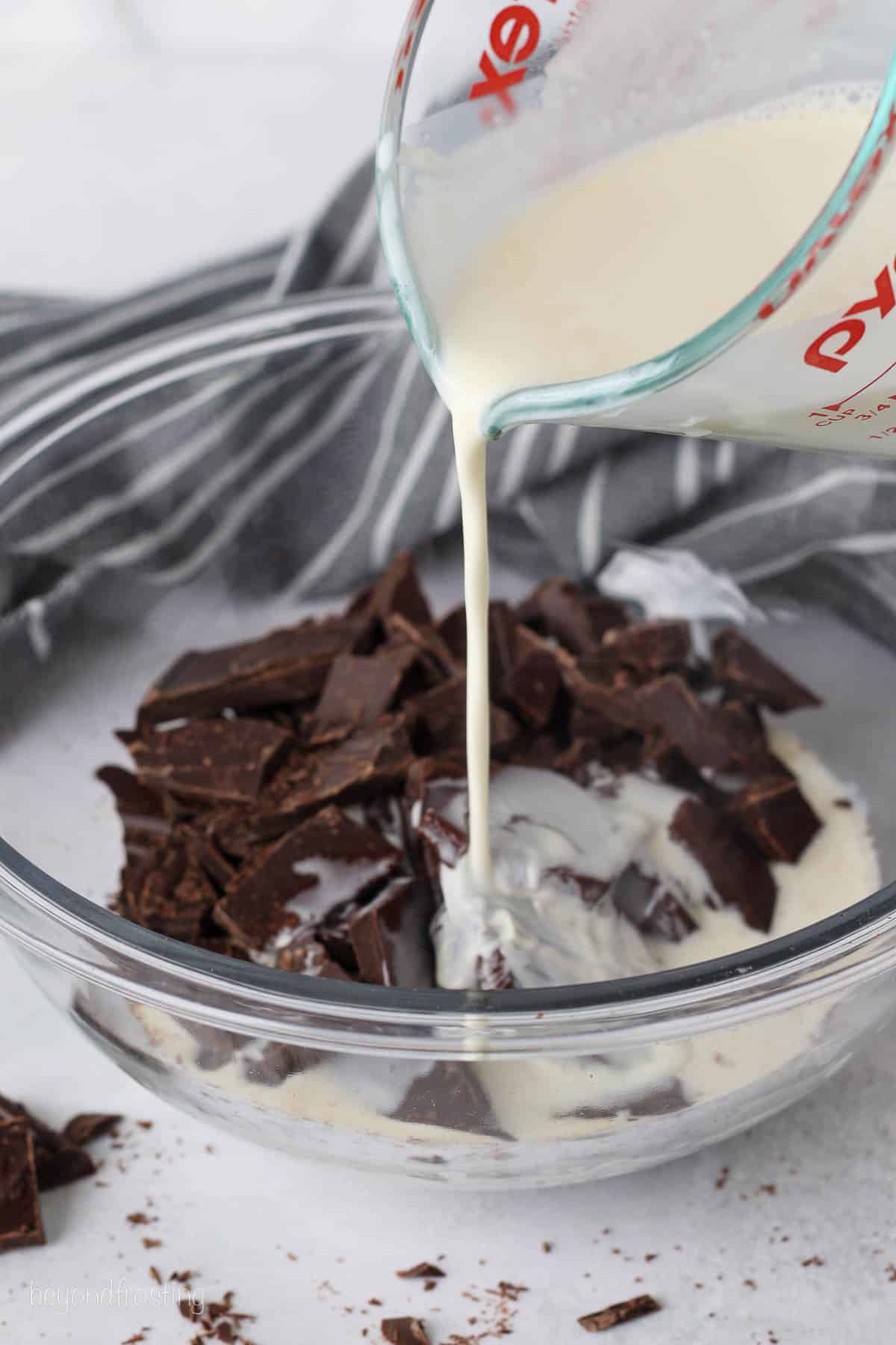 Heated heavy cream is poured over chopped chocolate in a glass bowl.