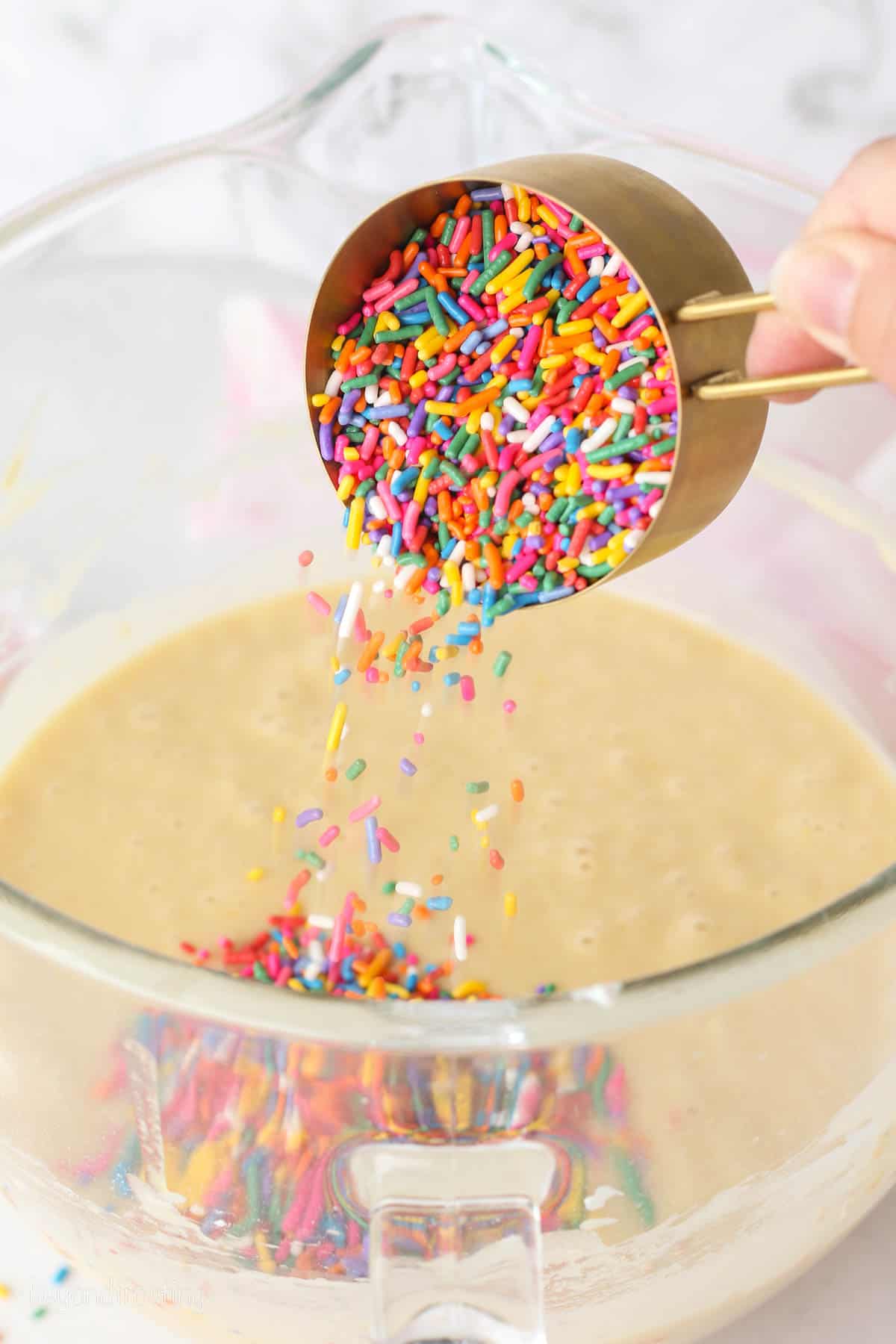 A measuring cup of rainbow sprinkles added to cake batter in a glass bowl.